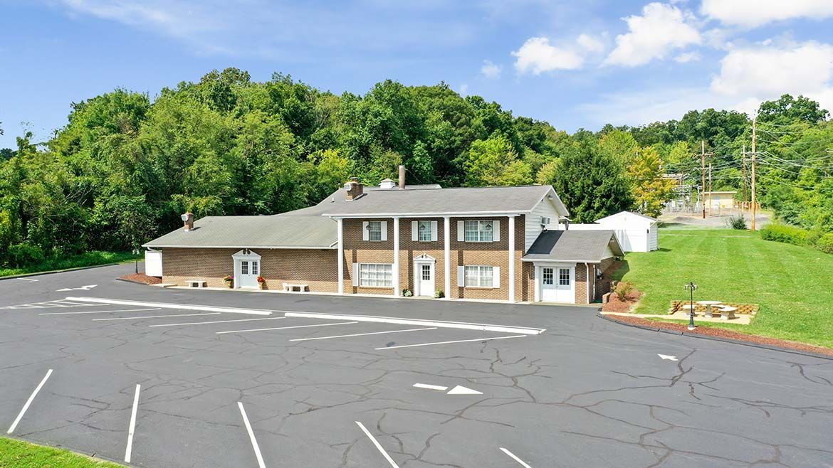 Small beige building with a gray roof beside a large empty parking lot and green hillside.