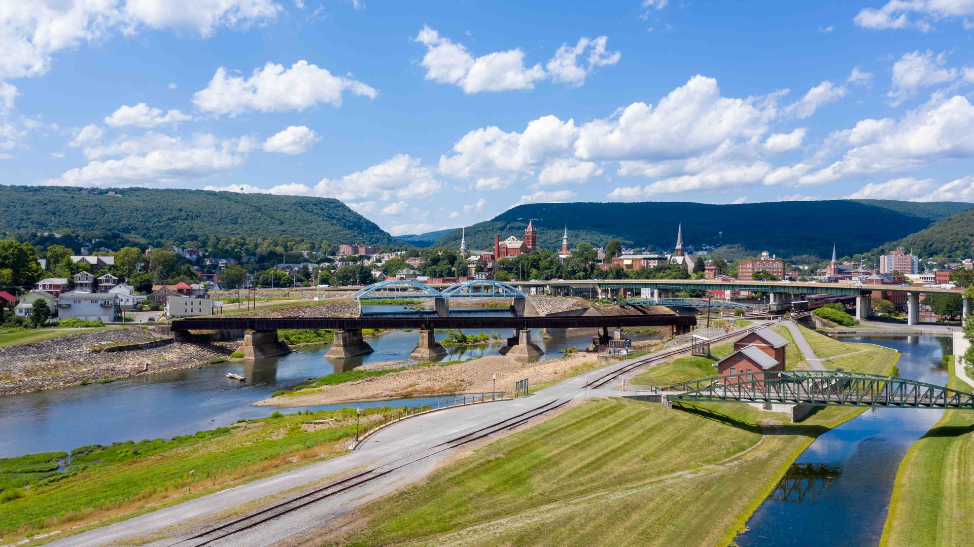Wide river valley town with a bridge, roads, green fields, and hills under a partly cloudy sky