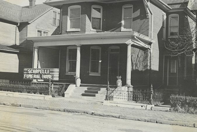 Black-and-white photo of a two-story house with a funeral home sign on the porch in a residential street