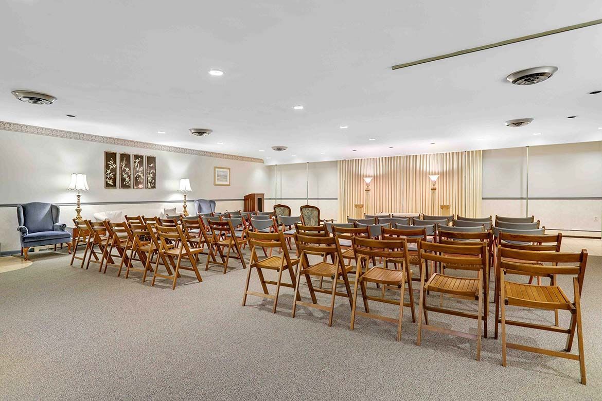 Empty community room with rows of wooden chairs facing the front and beige curtains at the windows.