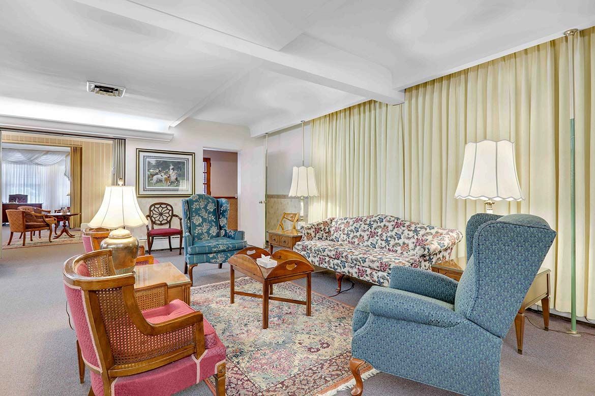 Bright living room with patterned sofas, armchairs, lamps, and curtains on carpeted floor.