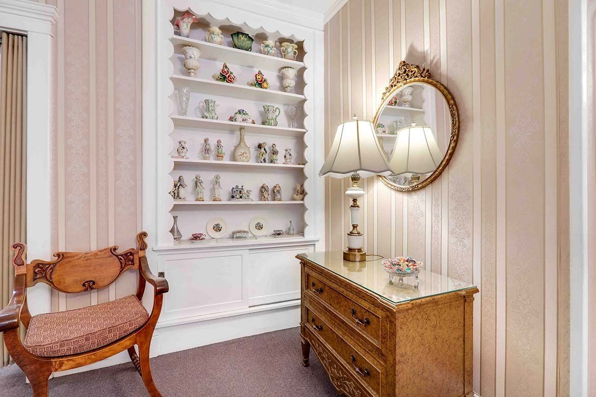 Elegant corner with white built-in shelves of figurines, ornate mirror, lamp, and antique chair and dresser