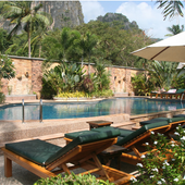 Poolside lounge chairs beside a tropical resort pool with stone wall and greenery.