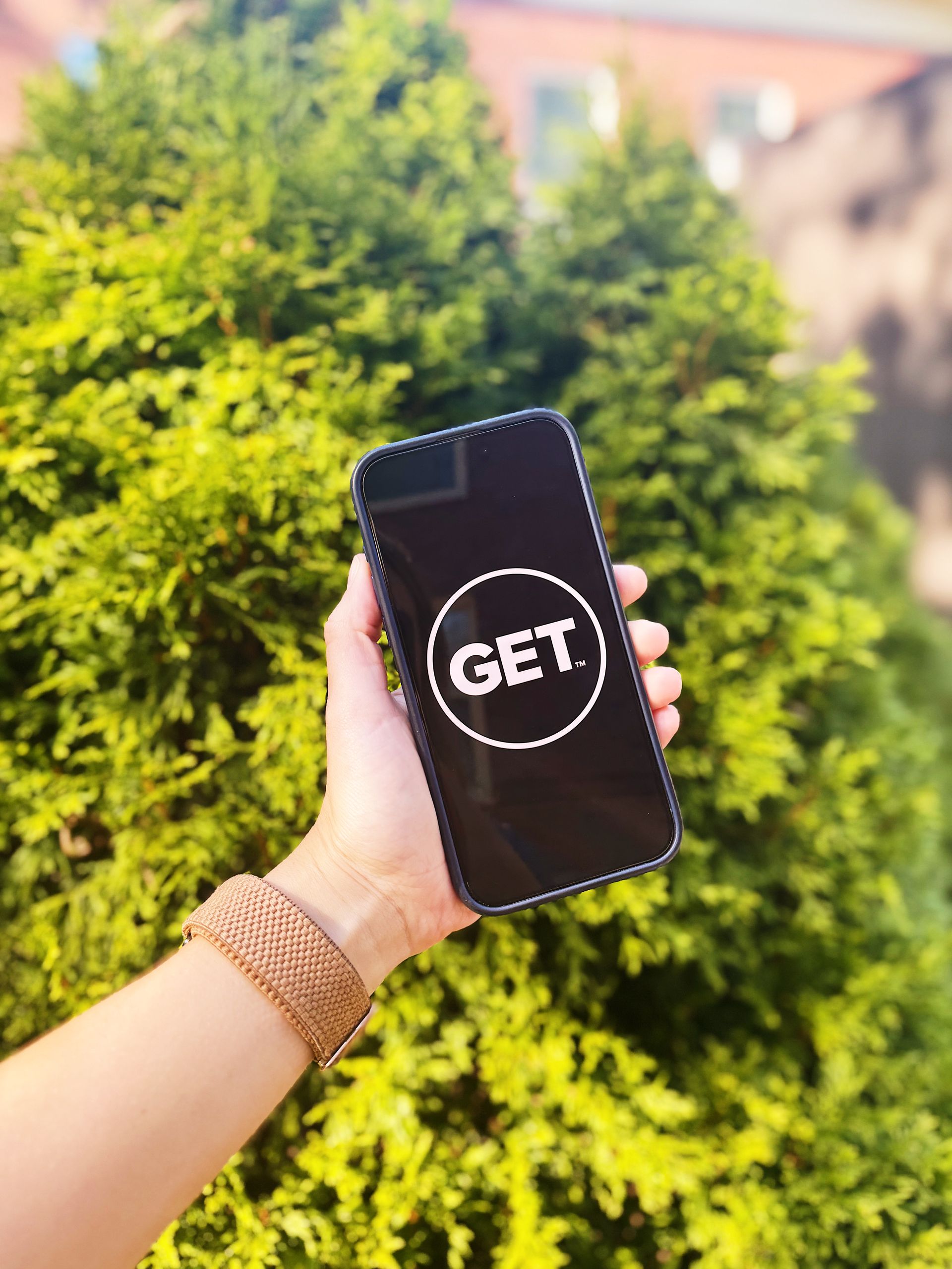 Hand holding a phone displaying a GET app logo against a backdrop of green foliage.
