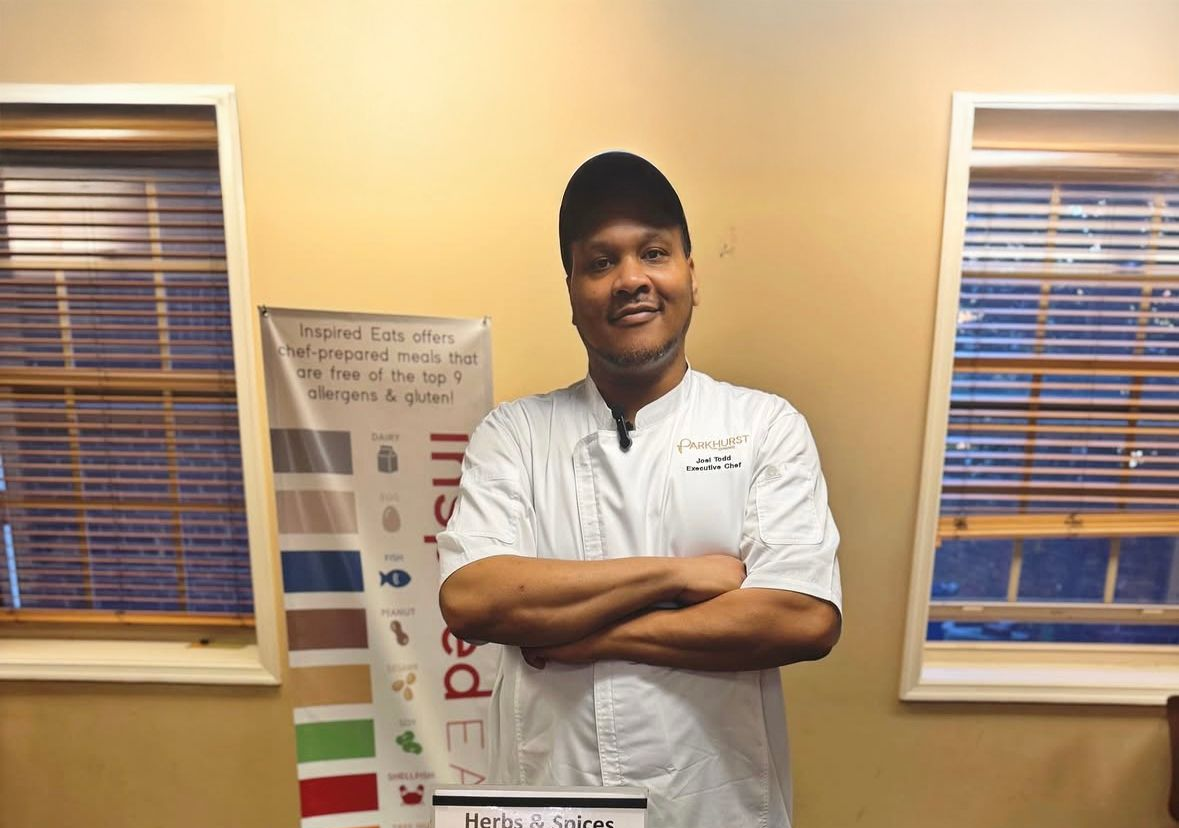 Chef stands at a table with herbs and spices. He's smiling with arms crossed, in front of a banner.