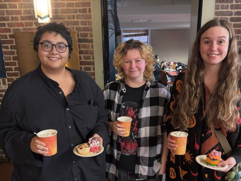 Three people stand smiling indoors, holding orange cups and small treats, in front of a brick wall.