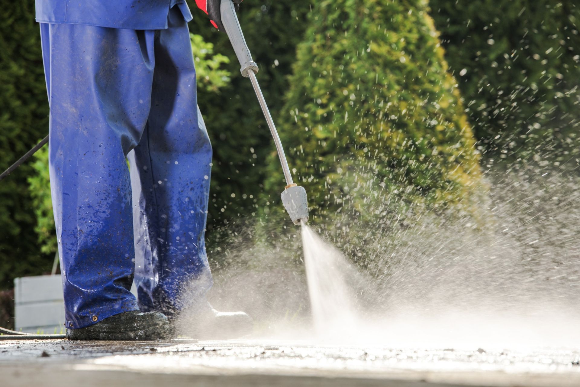 Person in blue overalls power washing a concrete surface with spray of water.