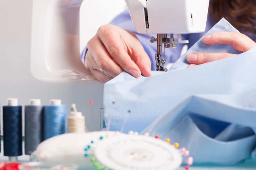 Persona che cuce un tessuto azzurro con una macchina da cucire; sono visibili rocchetti di filo e spilli.