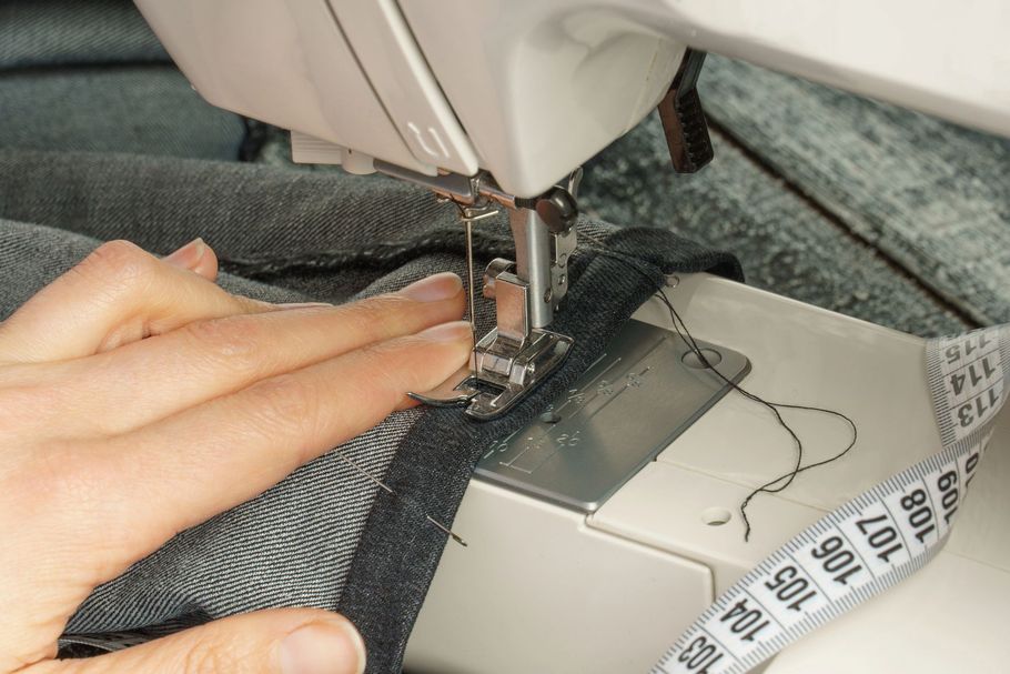 Mani di una persona che cuce tessuto denim su una macchina da cucire bianca. Un metro a nastro sulla destra.