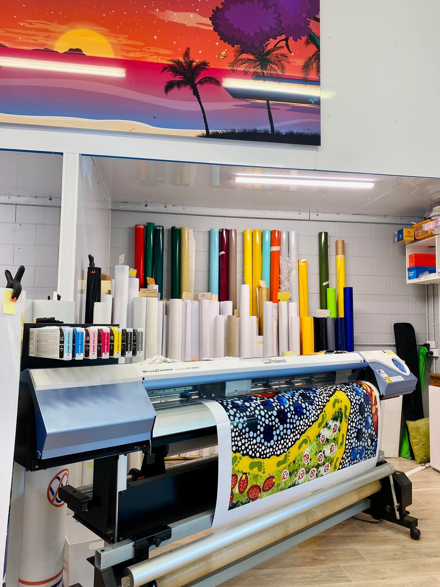 A large format digital printer in a workshop, with various colorful vinyl rolls stored on shelves and a sunset wall art.