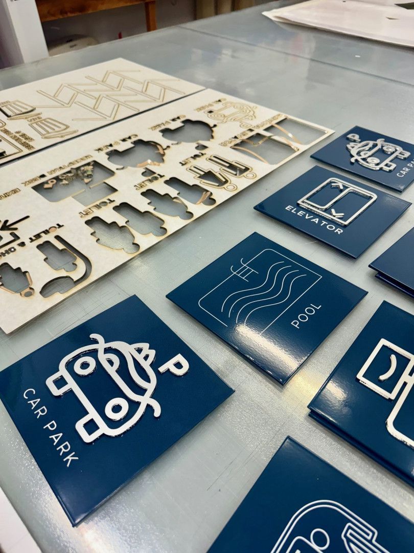 Laser-cut wooden shapes and dark blue signage tiles with white icons for wayfinding, including Pool, Car Park, and Elevator.