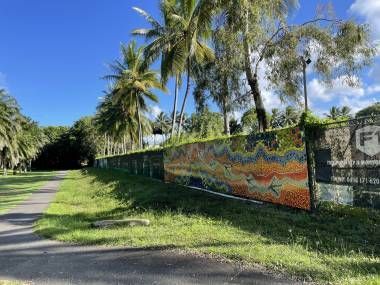 A paved path runs beside a long construction fence decorated with a colorful, textured mosaic mural, lined by palm trees.