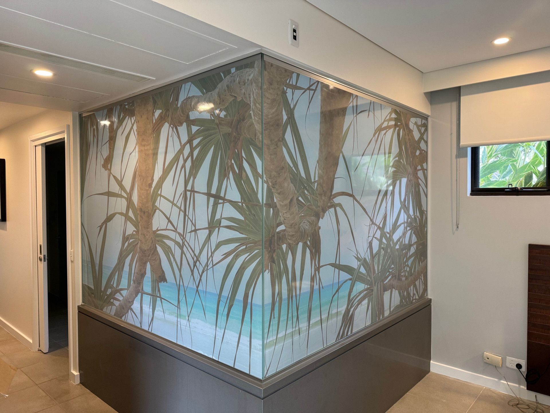 A glass corner partition features a semi-transparent, tropical beach mural with palm trees over turquoise water.