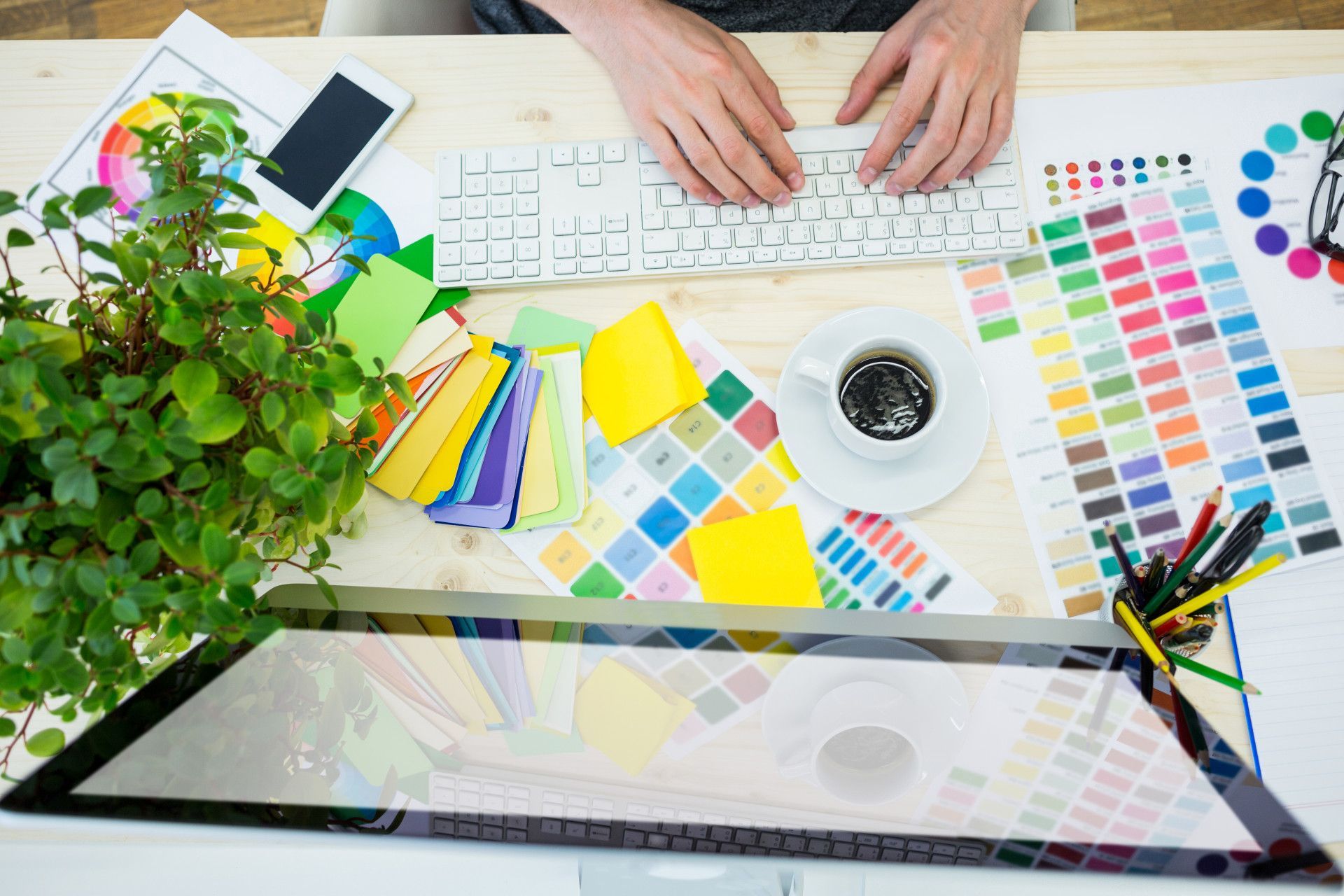 A person types on a keyboard surrounded by color swatches, a plant, a coffee cup, and a monitor on a wooden desk.