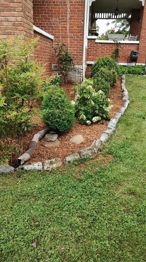 Small landscaped garden bed with stones, mulch, shrubs, and a brick house wall nearby.
