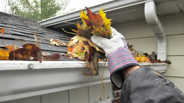 Gloved hand clearing wet autumn leaves from a rain gutter beside a house roof