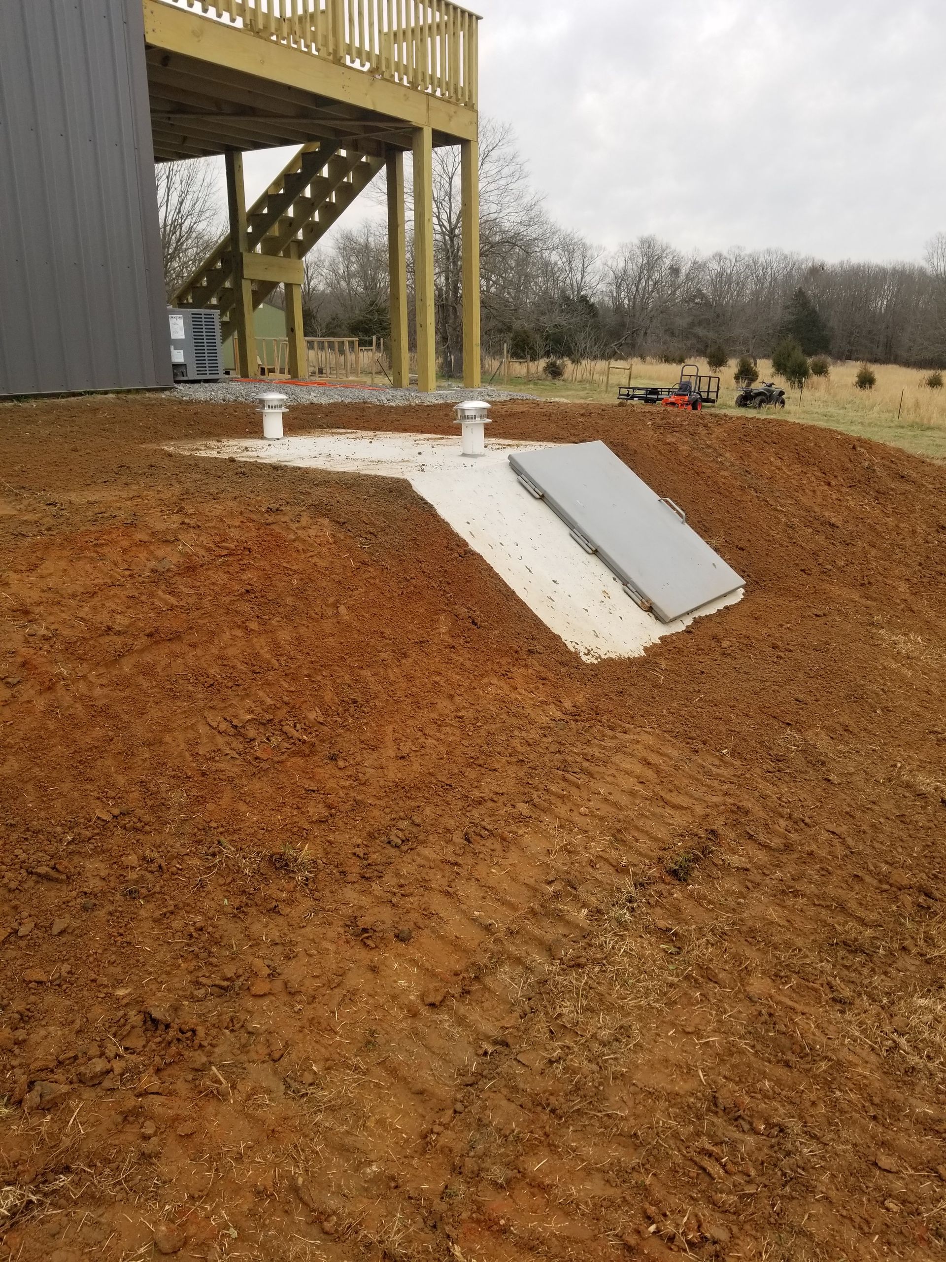 Exterior view of a building with a buried storm shelter entrance and a wooden deck. The ground is reddish-brown dirt.