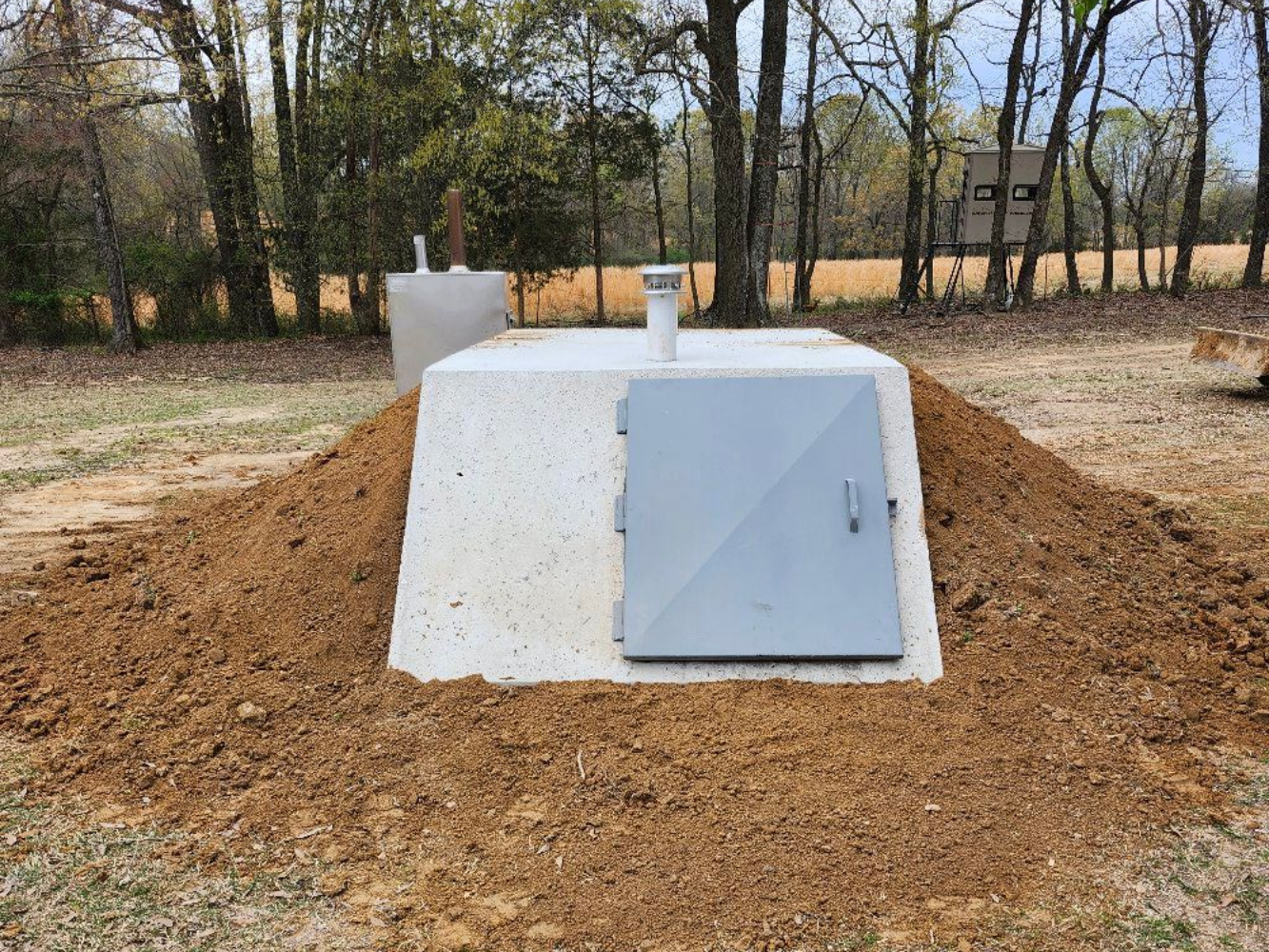 Concrete bunker with gray door, covered in dirt, in grassy field with trees.