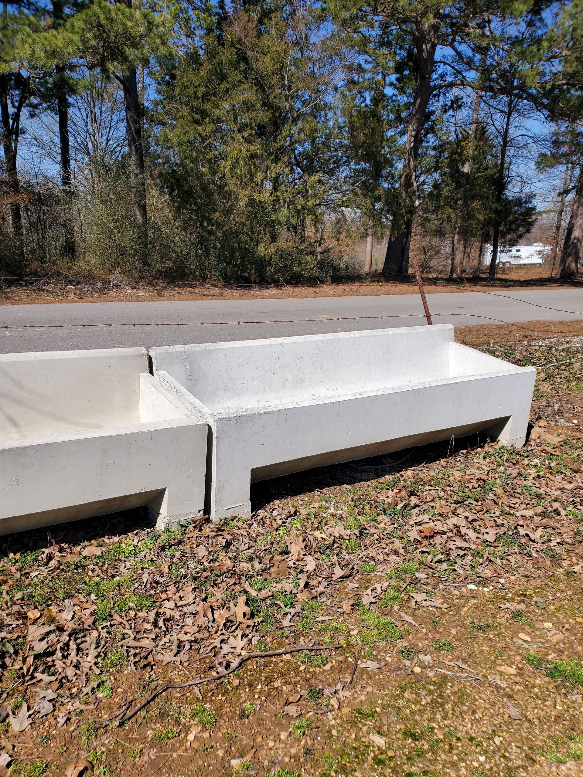 Two gray concrete troughs sit on grass next to a road, with trees in the background.