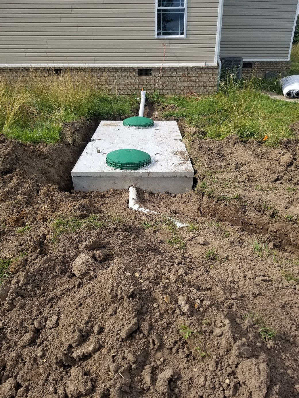 Concrete septic tank with two green lids in a dirt excavation, near a building.
