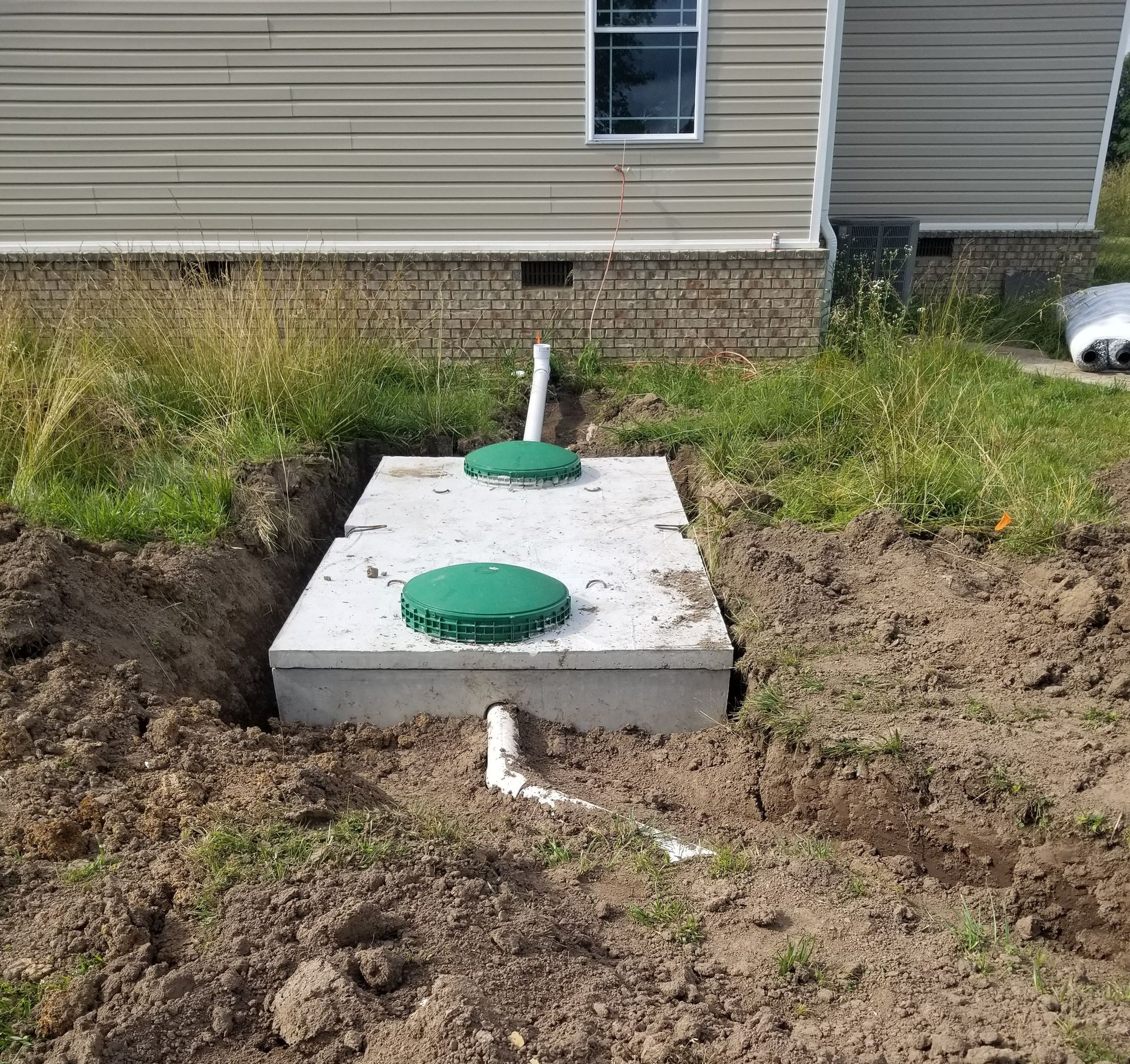 Septic tank installed near a house, with two green lids and connecting pipes in a trench.
