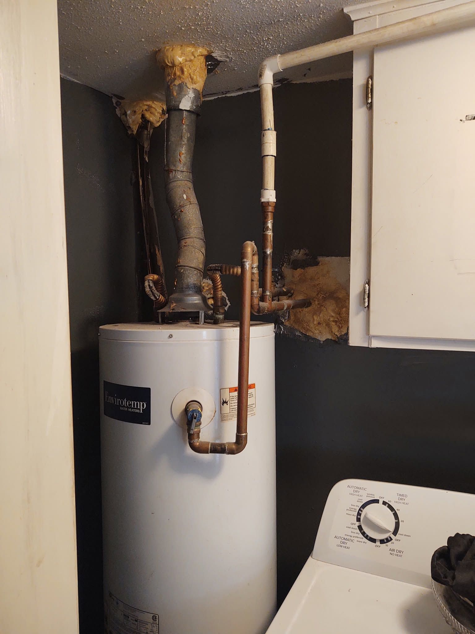 Water heater with exposed pipes in a cramped utility closet, beside a washer and cabinet.