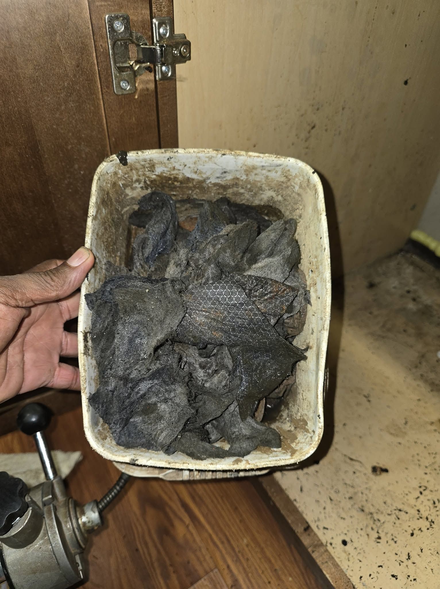 Hand holding a container filled with dark gray ash beside a wooden cabinet and floor debris