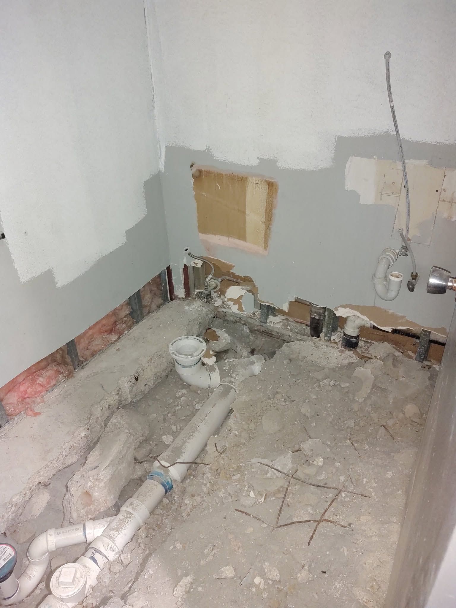 Damaged bathroom under renovation, with exposed pipes, broken wall plaster, and debris on the floor