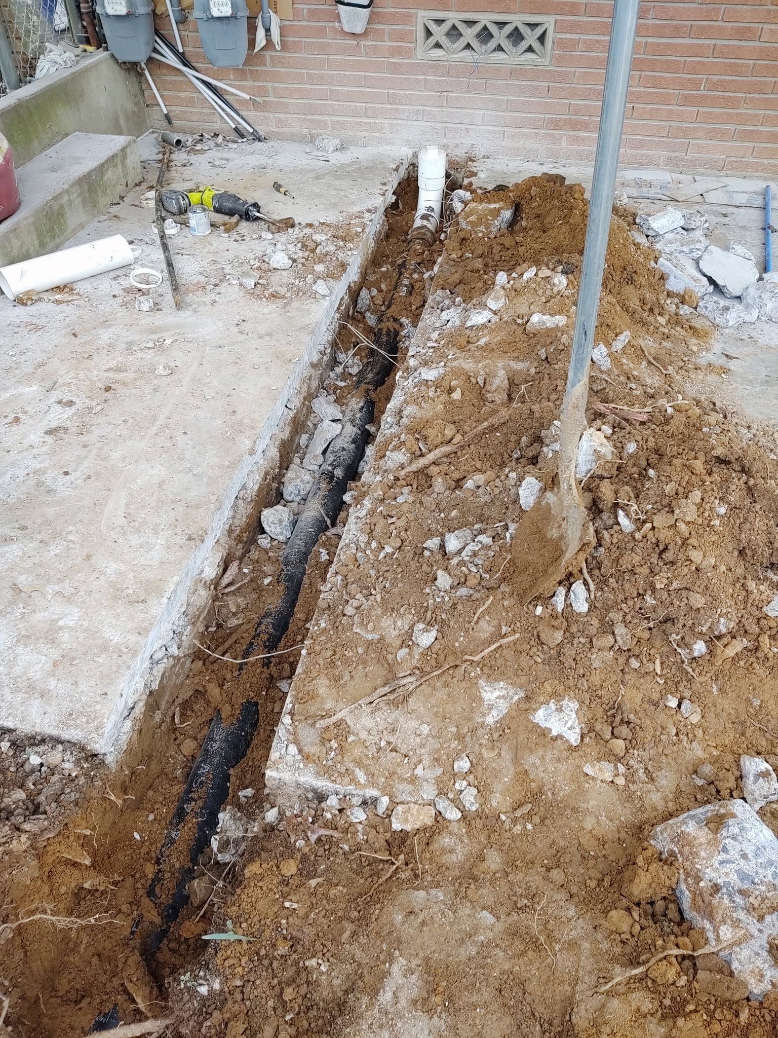 Trench dug through a dirt yard beside a brick wall, with exposed pipes and construction debris.
