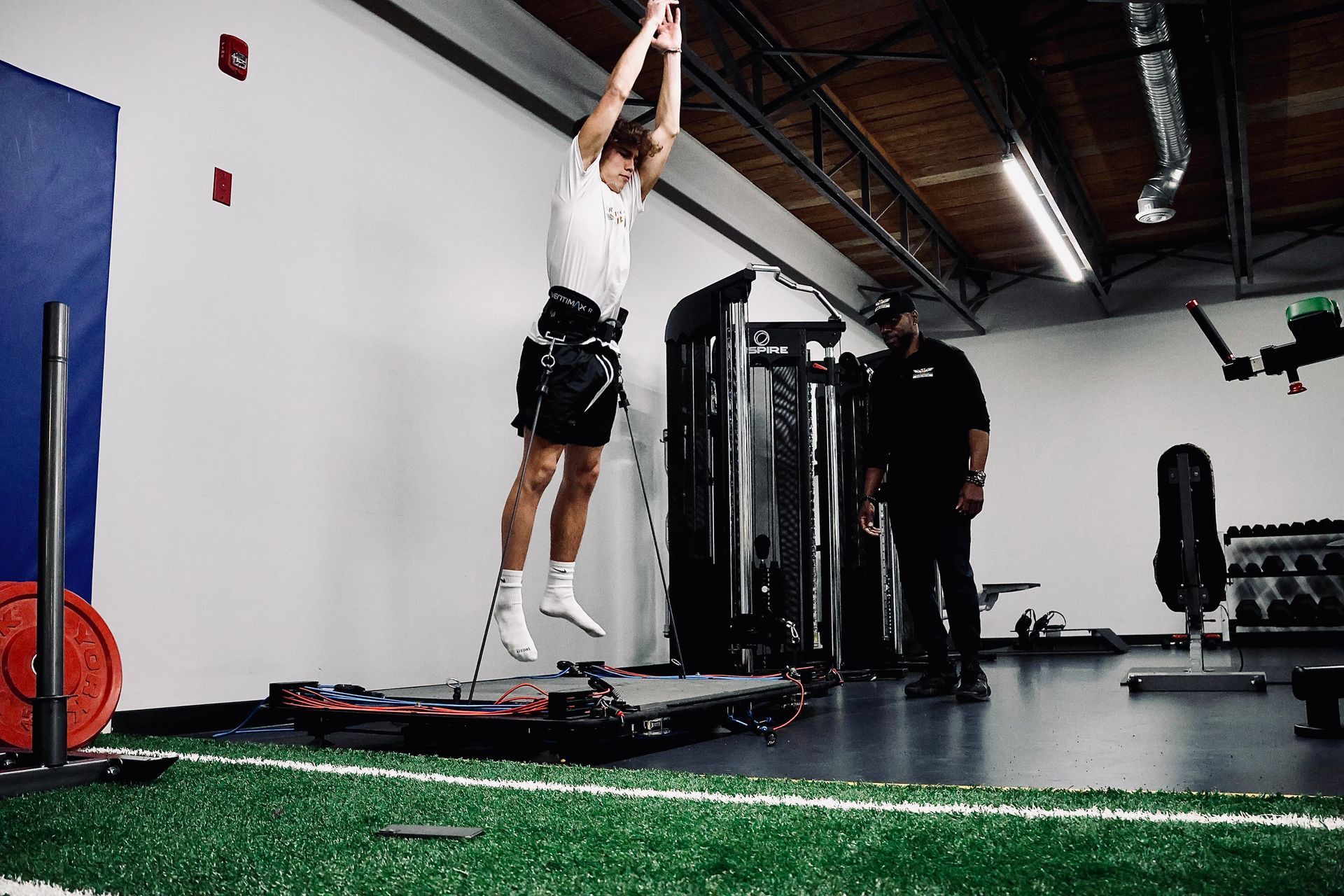 Man jumping high on a machine with a trainer, inside a gym with turf flooring.