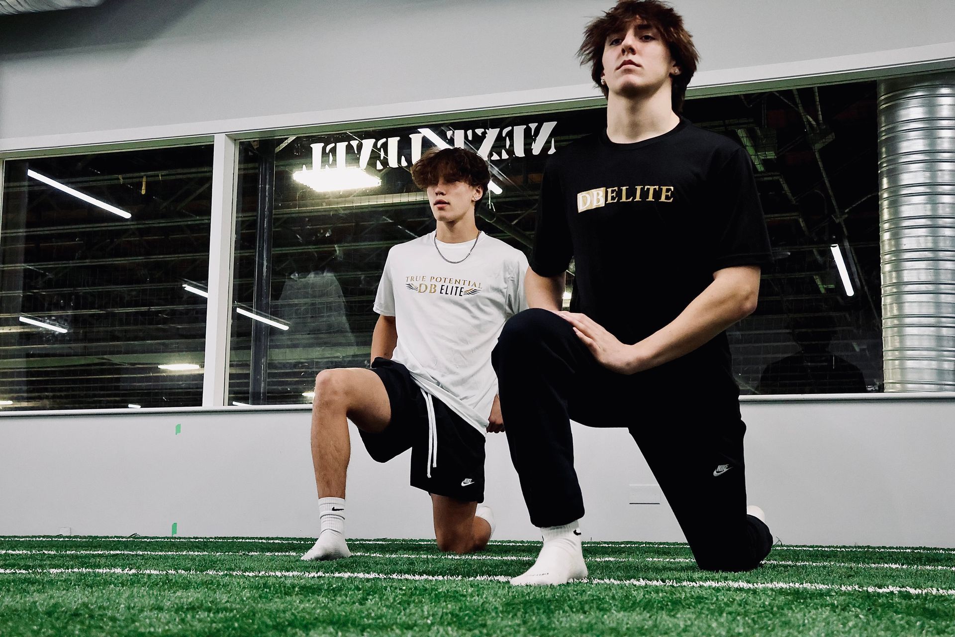 Two people kneeling on artificial turf, stretching in a gym with windows. One wears a black shirt, the other a white shirt.