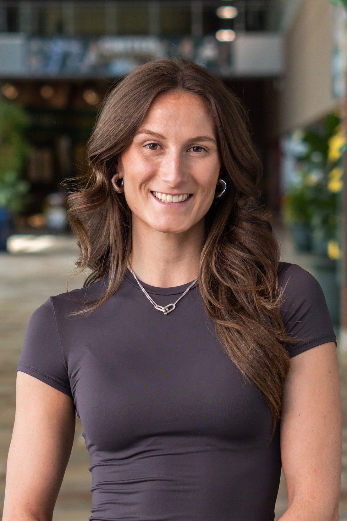 Smiling woman with long brown hair in a dark gray top, standing outdoors with a blurred background