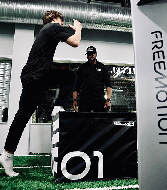 A person jumps onto a black box in a gym, watched by an instructor.