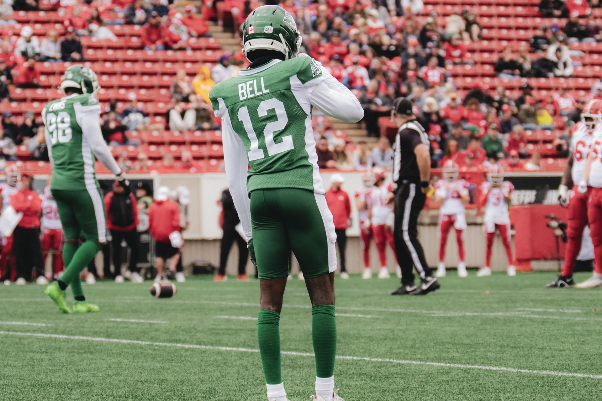 Football player in green jersey #12 stands on the field during a game, with crowd and sideline in the background