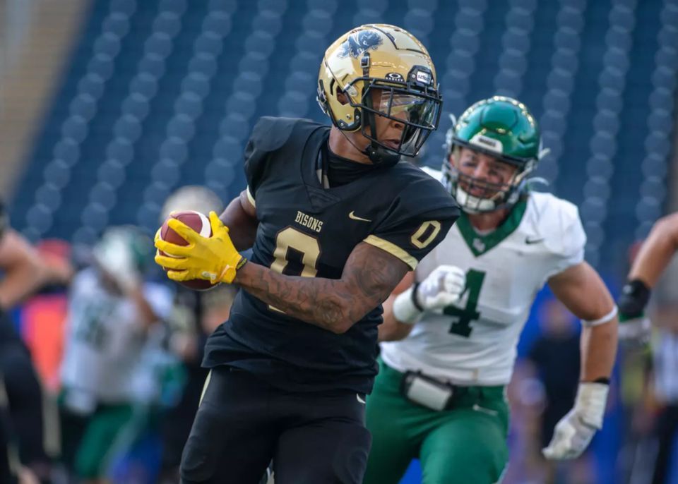 Football player in black and gold runs with the ball while a defender in green chases behind