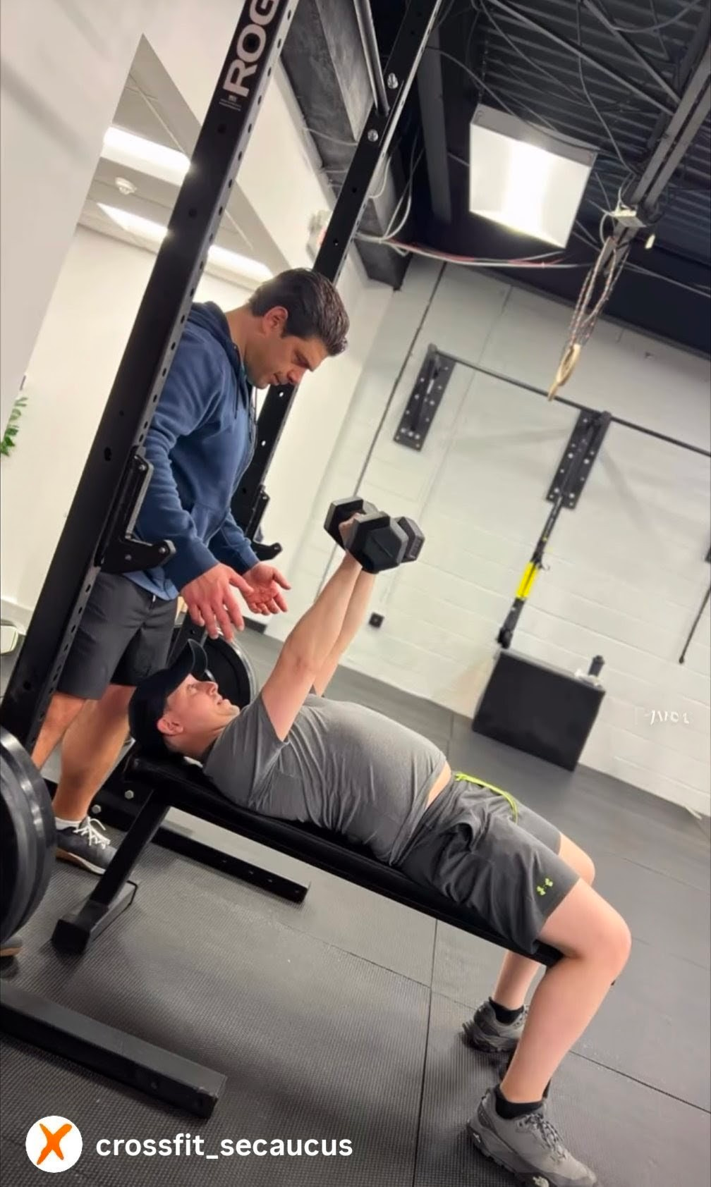 Two people training with dumbbells in a CrossFit gym, one spotting the other on a bench press.