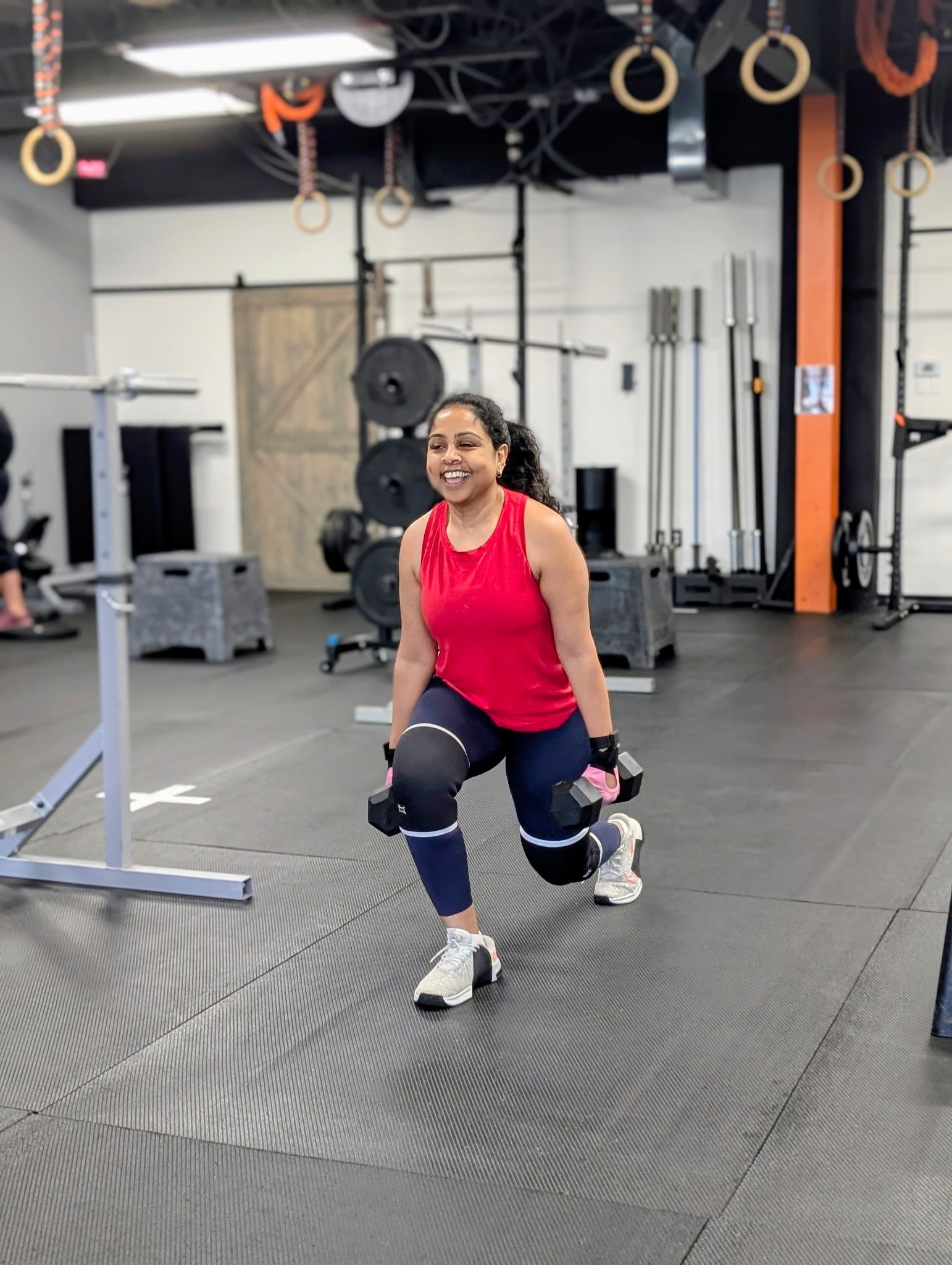 Woman in red top doing lunges in a gym with weight equipment and rings overhead