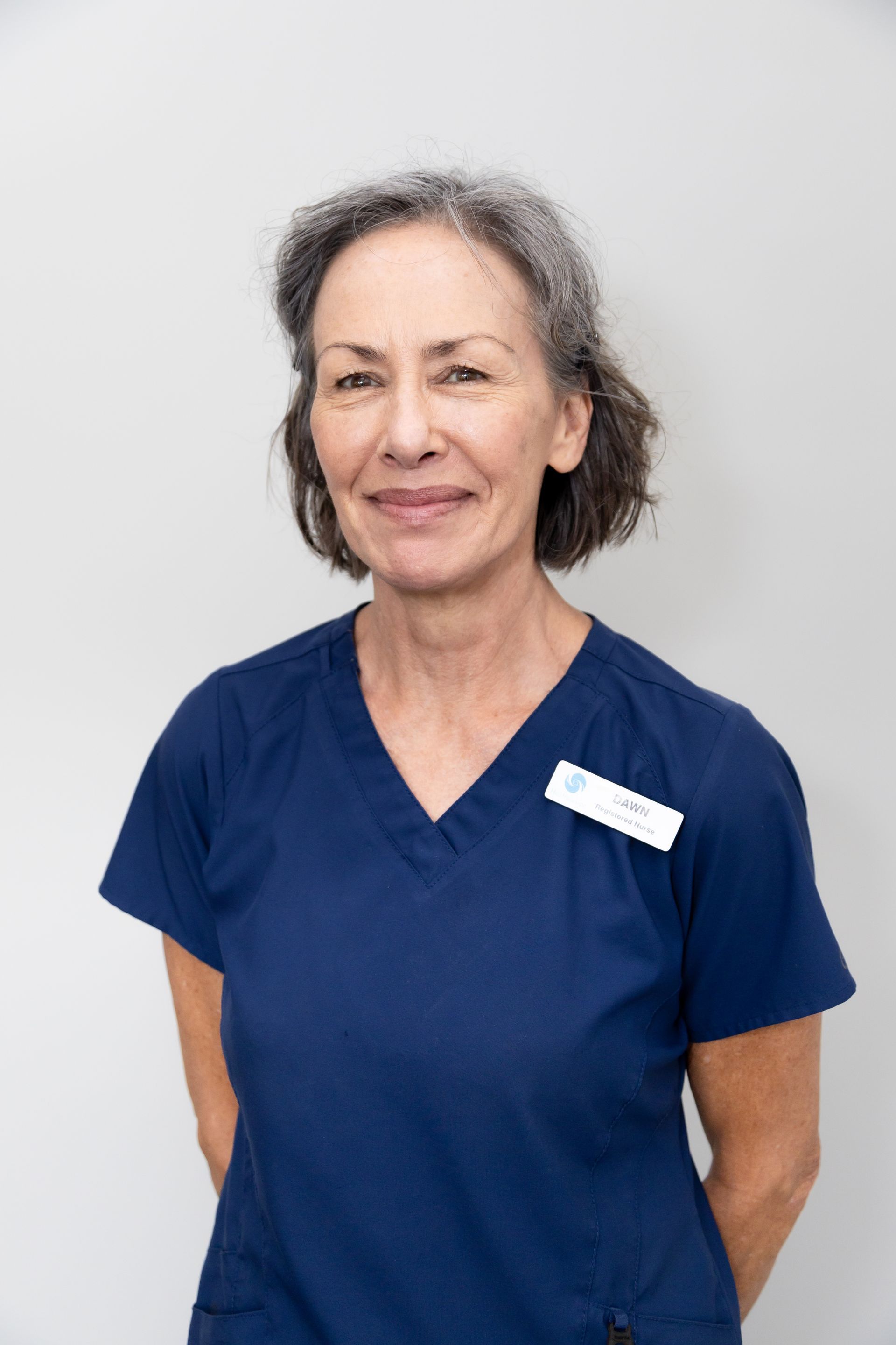 Woman in black scrubs smiles at the camera, arms behind her back, against a white wall.
