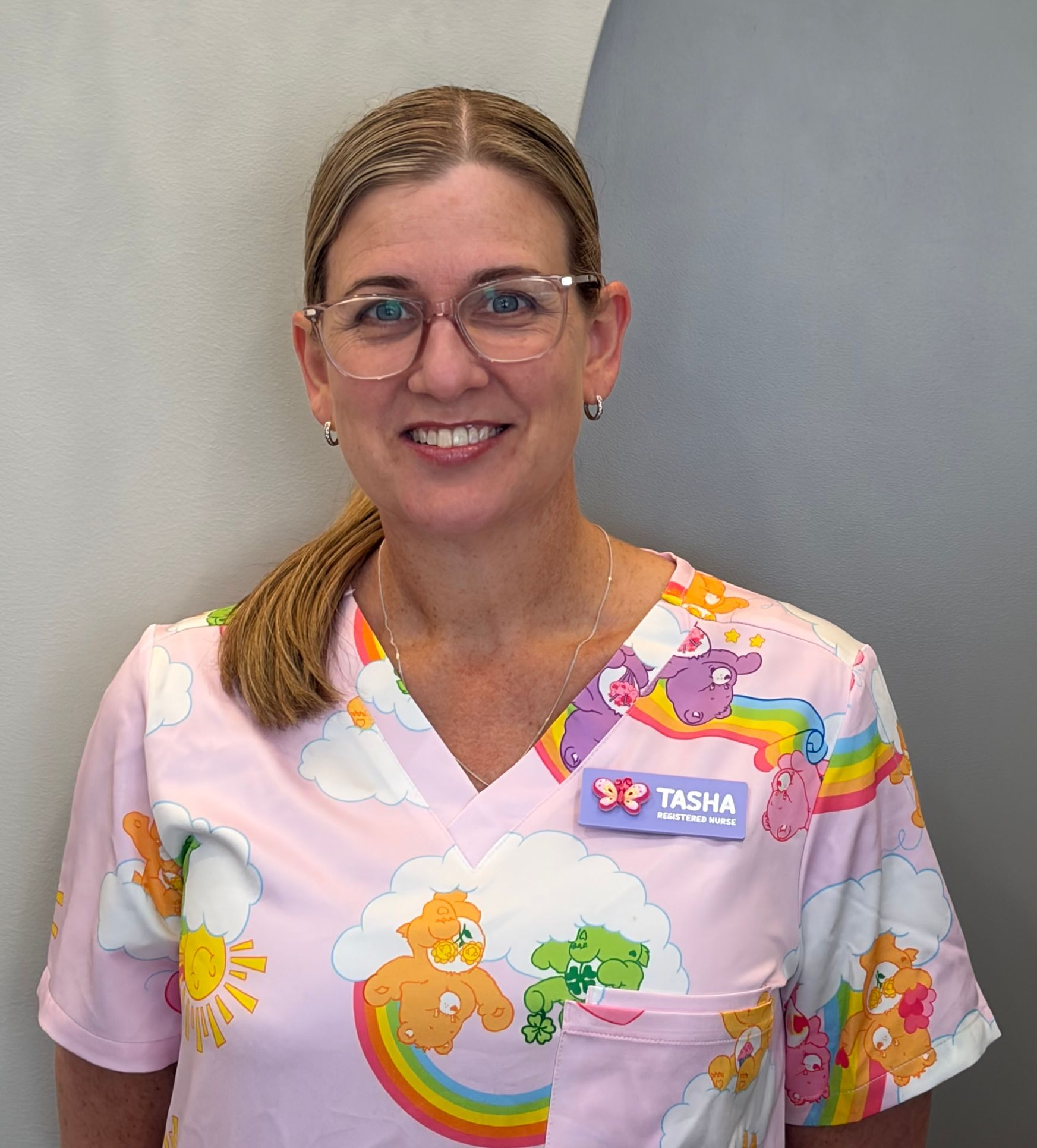 Woman in navy blue scrubs smiling, with name tag, against a white wall.