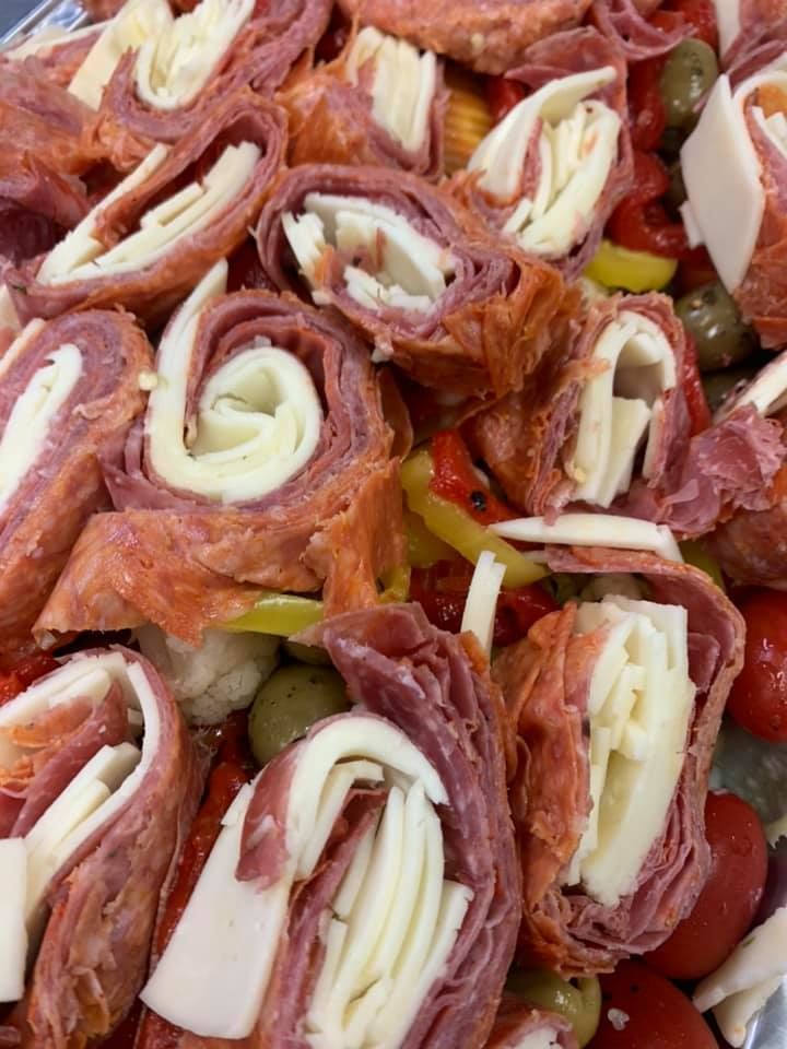 Rolled cured meat and cheese slices arranged over pickles, with red peppers in the background
