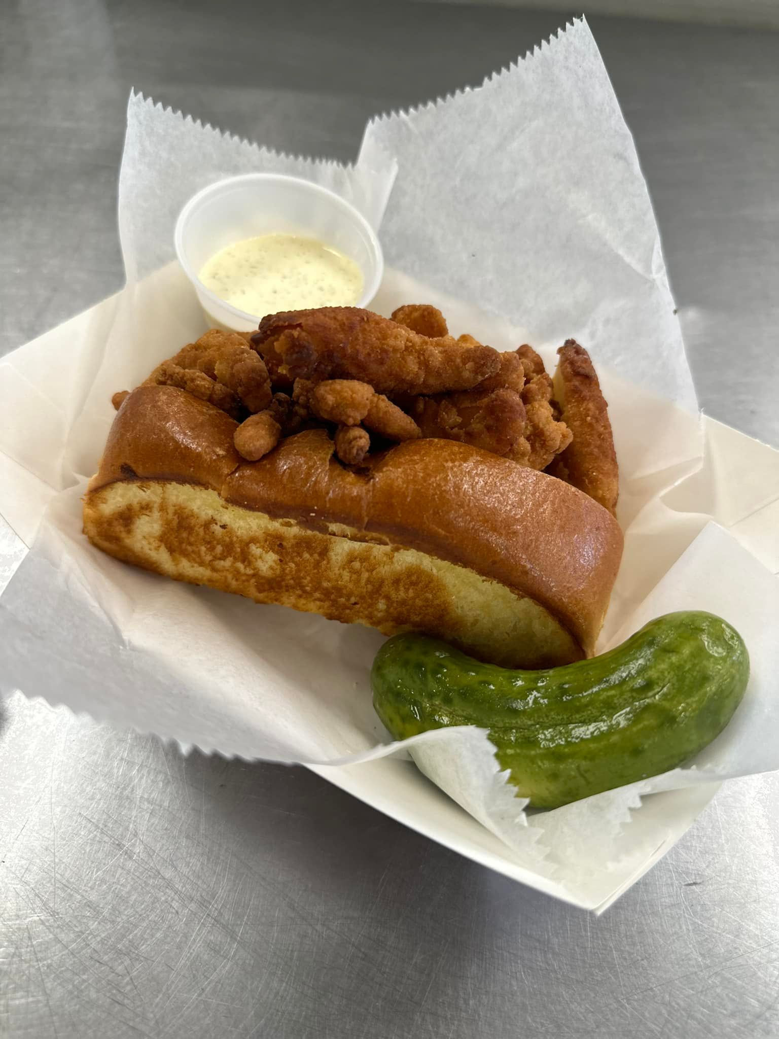 Fried sausage on a bun with mustard, served in a paper tray with a pickle spear and dipping sauce.