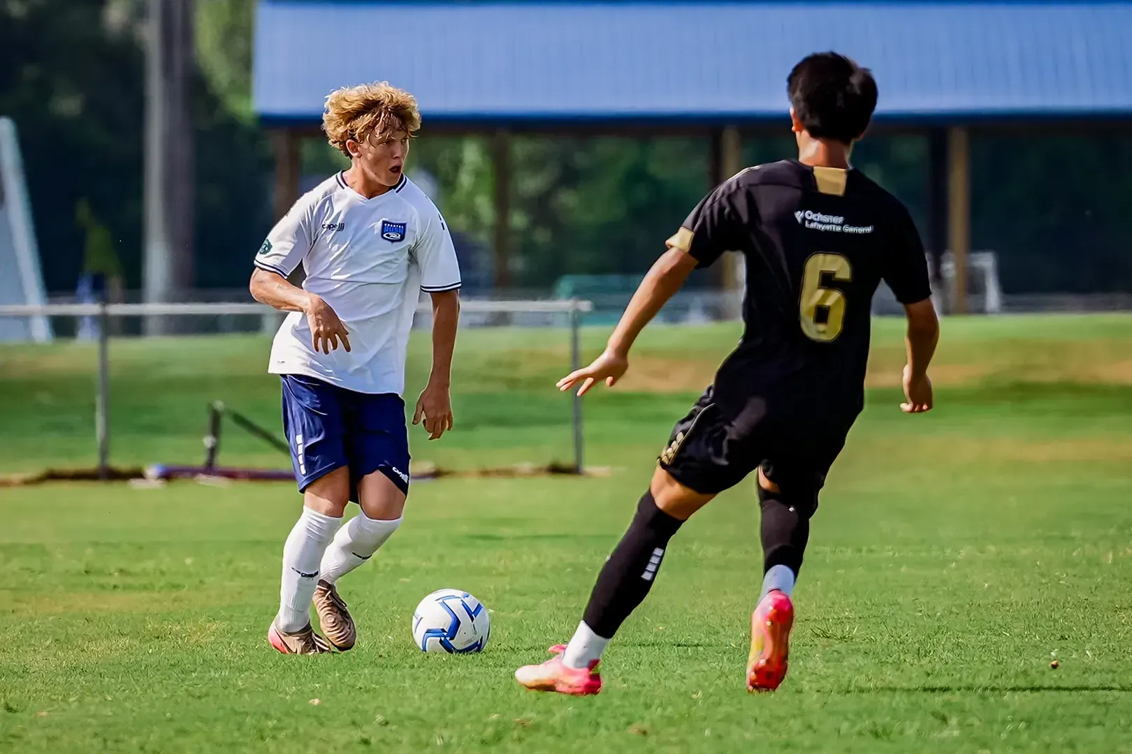 Two soccer players on a green field. One in white and blue, the other in black, about to kick the ball.