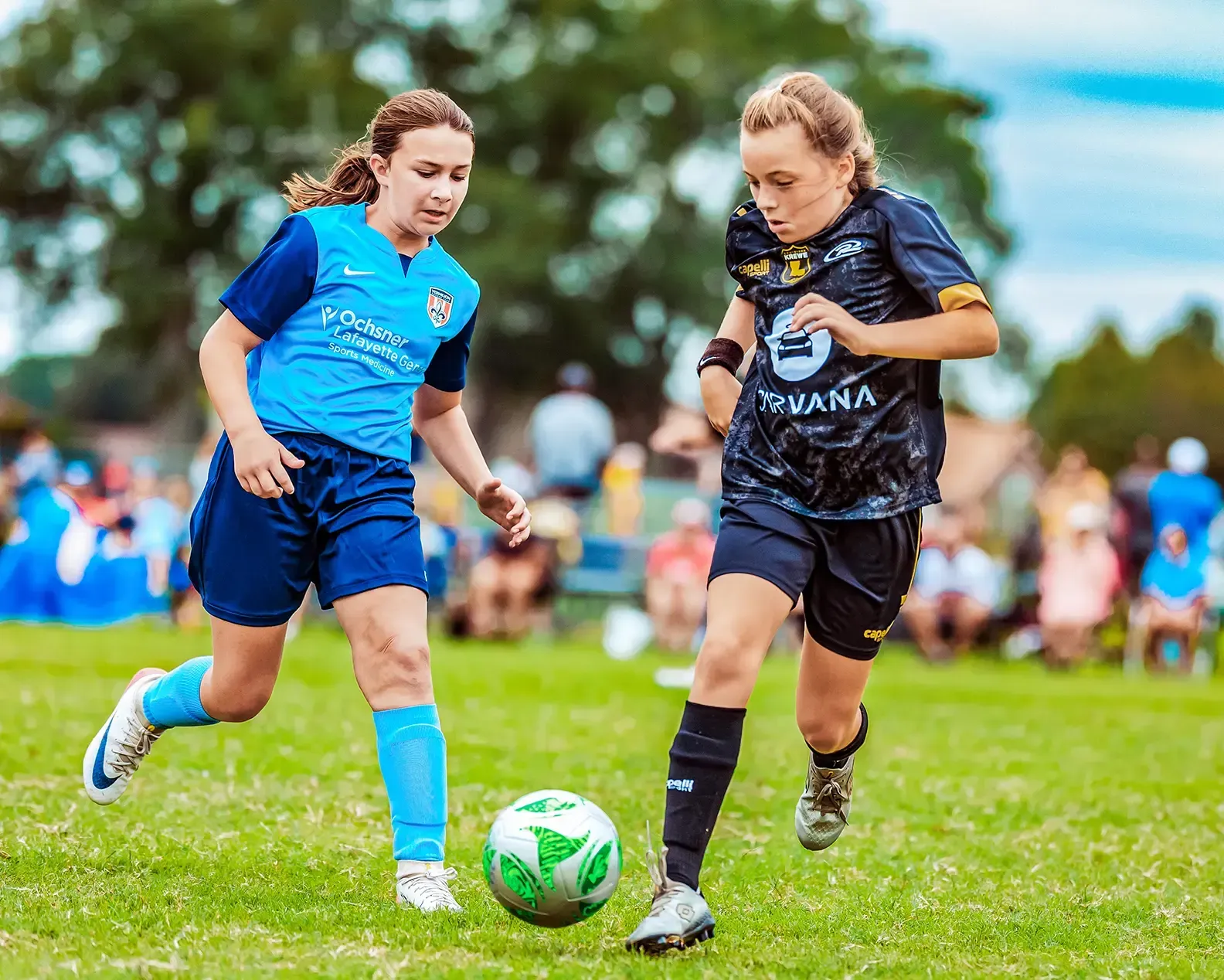 Two soccer players in action, chasing the ball on a green field. One in blue, one in black, both focused.