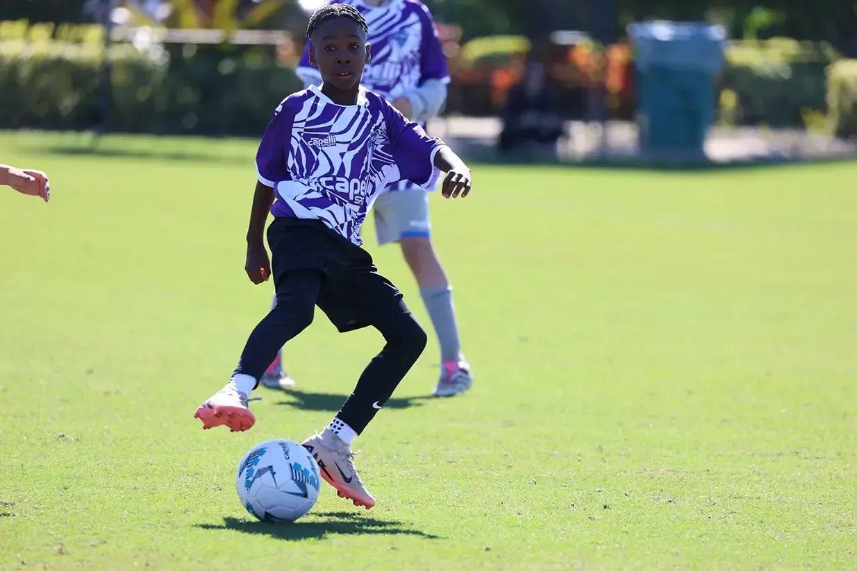 Boy in purple jersey dribbles soccer ball on green field.