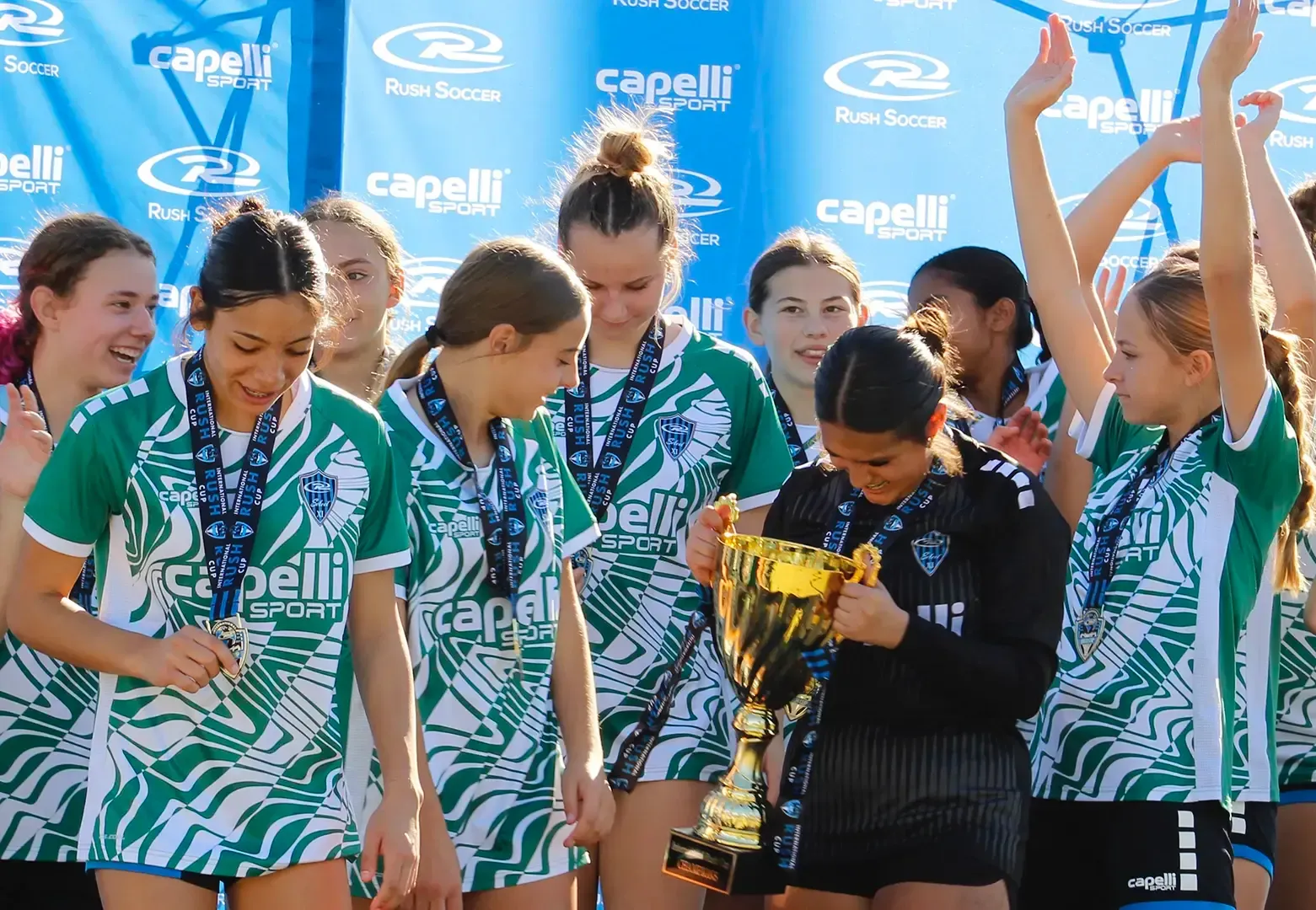 Girls' soccer team in green and white uniforms celebrating a win, one holding a trophy, others with medals.