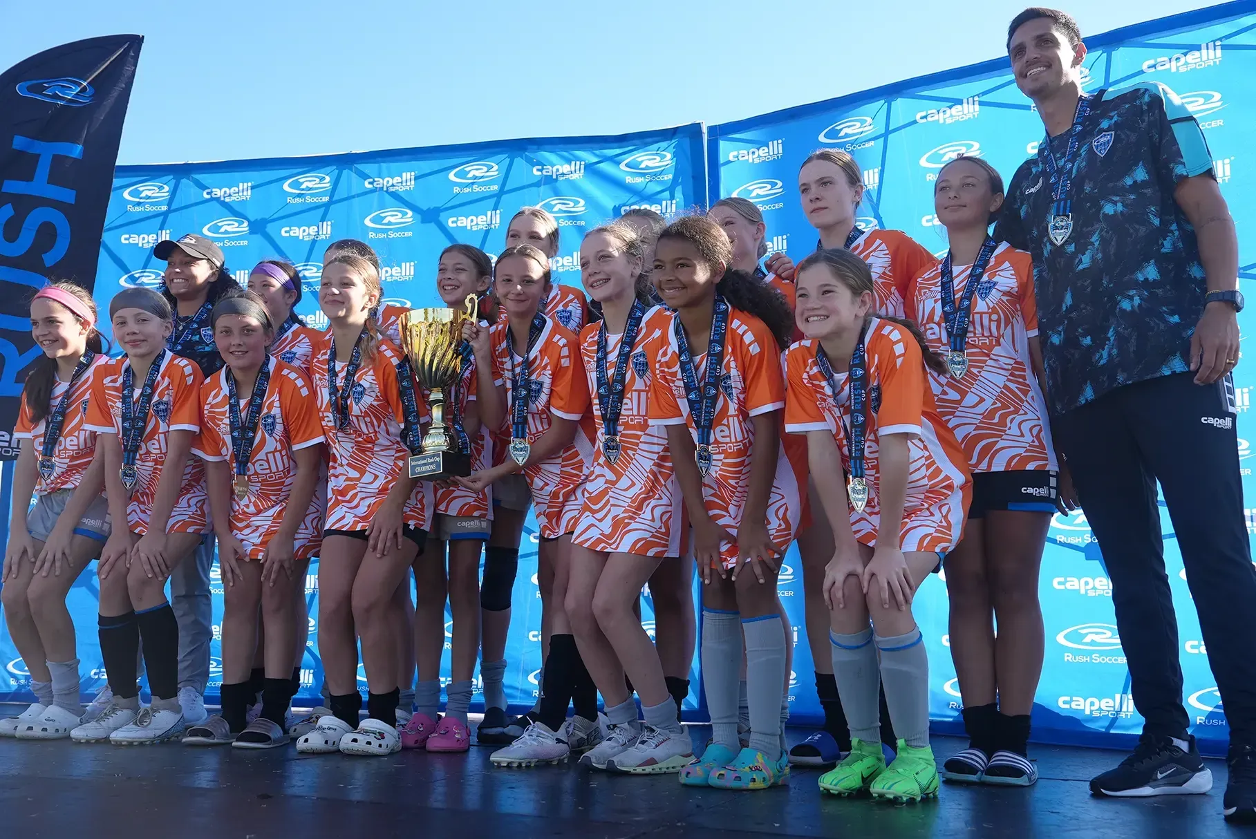 Youth soccer team in orange jerseys, holding trophy, smiling on podium. Blue backdrop, sunny outdoor setting.