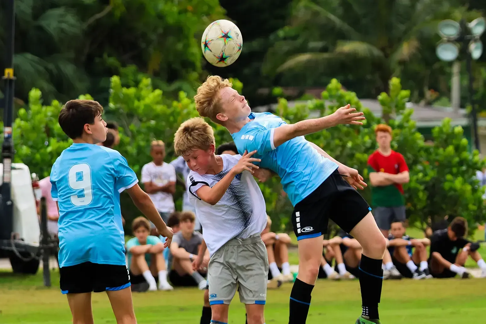 Soccer players in blue and white jerseys compete for a header on a green field. Spectators watch nearby.