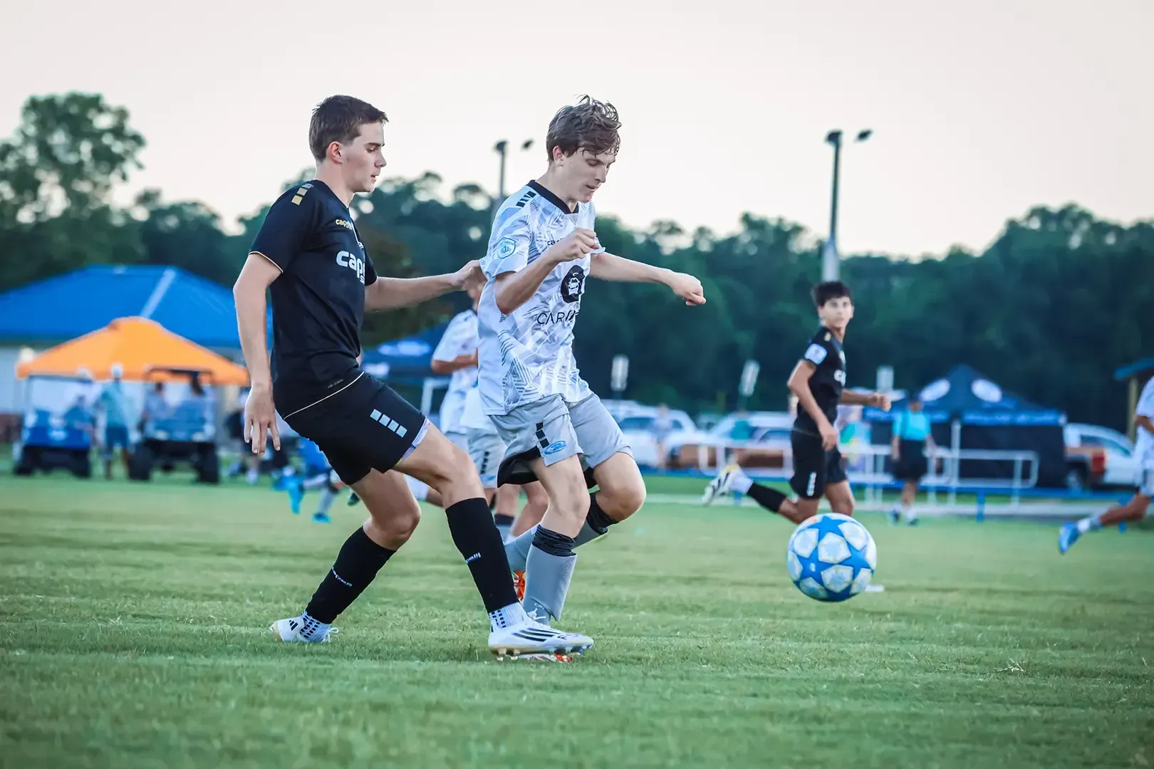Two soccer players on a field; one in black tries to block the other in white, who dribbles the ball.