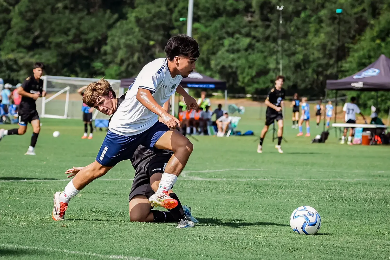 Soccer players compete on a green field. One player dribbles the ball while another dives to tackle.