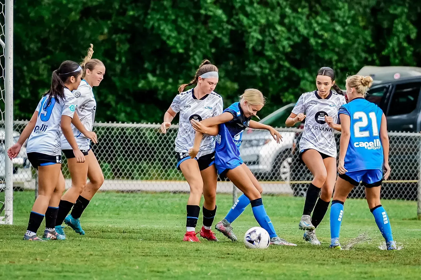 Soccer players in action on a green field. Two teams, one in blue, one in white. A player kicks the ball.